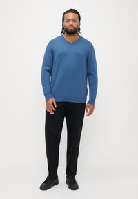 Niebieski sweter z dekoltem w serek, wykonany z miękkiego materiału, zestawiony z czarnymi spodniami i czarnymi trampkami. Na piersi niewielkie logo.