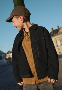 Chaqueta negra con costuras amarillas encima de una camisa marrón de botones. La chaqueta cuenta con un bolsillo y un corte holgado. Una gorra oscura completa el look.