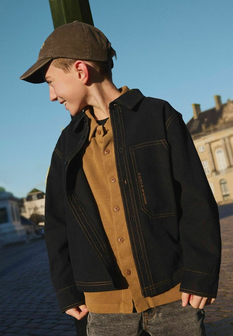 Chaqueta negra con costuras amarillas encima de una camisa marrón de botones. La chaqueta cuenta con un bolsillo y un corte holgado. Una gorra oscura completa el look.