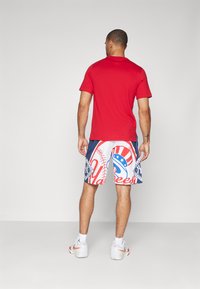 Camisa corta roja de manga corta combinada con pantalones cortos blancos que presentan un gráfico colorido con temática de béisbol y acentos azules. Zapatillas deportivas completan el conjunto.