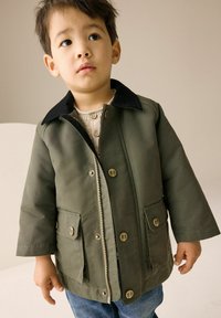 Grön jacka med dragkedja och svart krage, med två framfickor och bruna knappar. Bärs över en beige tröja. Denimbukser syns.