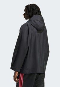 Svart huva jacka i vattentätt material, med vida ärmar, lös passform och en designdetalj längs vänster ärm.