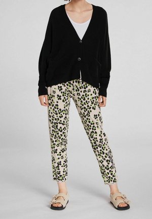 Person iført sort knappet cardigan, hvid top, beige bukser med grøn og sort blomsterprint og beige pelsede sandaler.