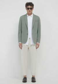 Blazer verde chiaro con rever a punta, camicia bianca, pantaloni crema e scarpe scure. Tessuto liscio con vestibilità su misura. Occhiali da sole a forma rettangolare.
