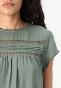 Frau trägt eine salbeigrüne Bluse mit Spitzen-Einsätzen, Falten unter der Brust und Pompon-Besatz am Ausschnitt und den Ärmeln.