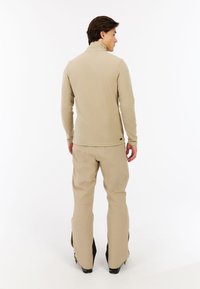 Beige fleece sweater med høj krave, struktureret stof, parret med beige udendørs bukser, der har elastisk talje og sidelommer.