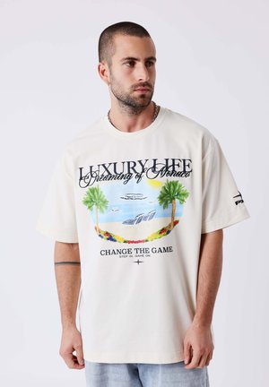 Hombre con pelo corto y barba que lleva una camiseta blanca extragrande con un gráfico de palmeras y escena de playa y el texto «Luxury Life, Dreaming of Monaco».