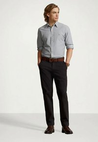 Ung man som står med händerna i fickorna, iklädd ljusgrå skjorta med knappar, mörka byxor, brun bälte, klocka och bruna skor, som tittar åt höger.