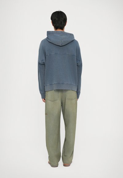 Felpa blu con cappuccio e cuciture a contrasto, abbinata a pantaloni in denim verde chiaro larghi con tasche posteriori.