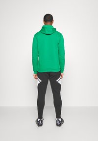 Grüne Kapuzenjacke mit Kordelzug-Haube, kombiniert mit schwarzen Sporthosen mit weißen seitlichen Akzenten. Die Schuhe sind schwarze Sportschuhe.