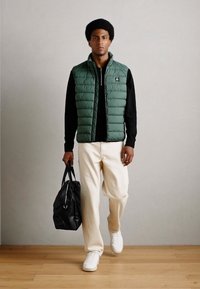 Grüne Puffweste über einem schwarzen Sweatshirt, cremefarbene Hose und weiße Sneaker; trägt eine schwarze Tasche. Minimalistisches Design mit einer glatten Textur.