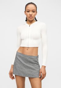 Jeune femme aux cheveux tressés portant un sweat à capuche court blanc zippé et une mini-jupe à carreaux noir et blanc, debout devant un fond uni.