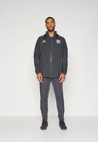 Veste de sport grise avec capuche et fermeture éclair, ornée d'accents dorés et du logo de l'équipe. Accompagnée d'un pantalon gris assorti et de chaussures de sport noires.