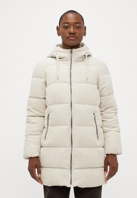 Beige cordfløjs pufferjakke med hætte, lodret quiltet mønster, to forreste lynlåslommer og en hel lynlås fortil.
