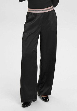 Frau trägt schwarze Hosen mit weitem Bein, gestreiftem elastischem Bund und schwarzen Absatzschuhen, vor schlichtem grauem Hintergrund.