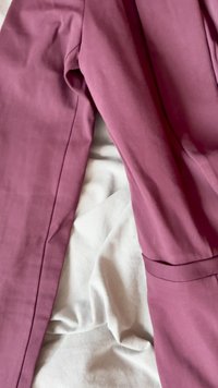 Fioletowa kurtka z gładkiego materiału o prostym kroju, z boczną kieszenią i szwami wzdłuż rękawów. Pod spodem złożony biały materiał.