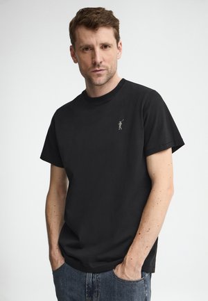 Mand med kort brunt hår iført en sort t-shirt med et lille golfspillerlogo på brystet, hænderne i lommerne, foran en ensfarvet hvid baggrund.