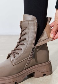 Botín de cuero marrón hasta el tobillo con cordones, cremallera lateral, forro de tela texturada y tacón grueso, con un puño acolchado y acentos en contraste.