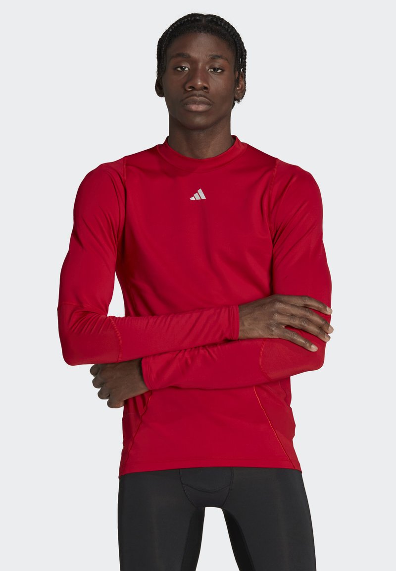 Rotes Langarm-Athletikshirt aus dehnbarem Material mit Rundhalsausschnitt; verfügt über ein reflektierendes graues Logo auf der linken Brust und Nahtakzente.
