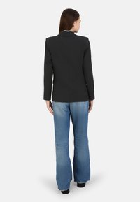 Blazer negro con un corte entallado, que presenta una abertura trasera, llevado sobre unos jeans acampanados de color azul claro y zapatos negros, mostrando la vista trasera.