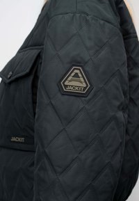 Black quilted jacket sleeve with a hexagonal patch featuring a stylized "A" and "JACK1T" logo, with a flap pocket in the background.