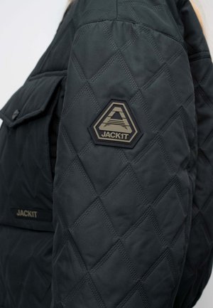 Mouw van een zwart gewatteerd jack met een zeshoekige patch met een gestileerde "A" en het "JACK1T"-logo, met daarachter een klepzak.