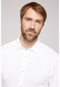 Camicia bianca con colletto classico e maniche corte, caratterizzata da una consistenza morbida e una leggera lucentezza. Il modello ha i capelli castano chiaro e la barba.