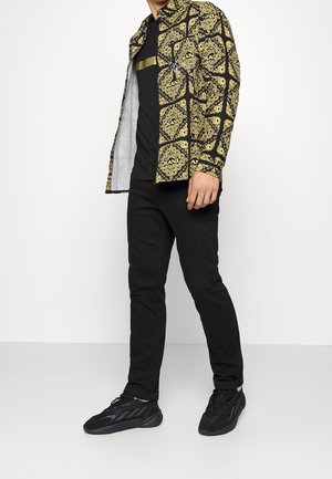 Man draagt zwarte broek, zwarte sneakers, zwart T-shirt en een open zwart-gouden patroon overhemd met het "KANI" logo.