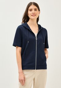 Hoodie zippé bleu marine à manches courtes, capuche et poches avant. Confectionné à partir d'un tissu doux avec une texture lisse. Équipé de quincaillerie argentée.