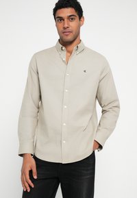 Camicia beige chiaro con bottoni, realizzata in cotone, con colletto classico, maniche lunghe e piccolo logo sul petto. Abbinata a jeans scuri.