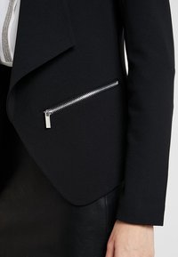 Svart blazer med en skarp design som har en diagonal framöppning. Inkluderar en ficka med dragkedja och detaljer i krom.