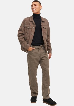 Mann lächelt, trägt eine braune Jeansjacke, schwarzen Rollkragenpullover, braune Hosen mit verstärkten Nähten und schwarze Schuhe, steht mit der Hand in der Tasche.