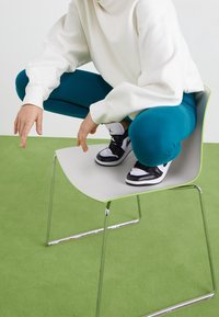 Vit oversized hoodie, teal leggings, svarta och vita sneakers, person i knästående på en minimalistisk grå stol med glänsande kromben på grön matta.