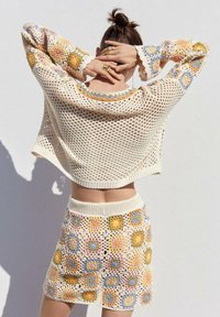 Kudotusta kahdesta osasta koostuva asu, johon kuuluu avoin neulekerroksellinen vaalean beige crop-top, jossa on värikkäitä kukkakoristeita, ja vastaava mini-hame, jossa on teksturoitu kuvio.