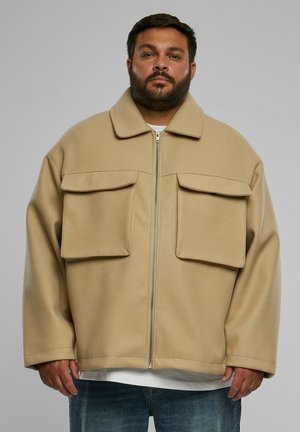 Beige oversized jacket with a collar, front zipper, and two large chest pockets. Material appears soft and textured. Worn over a white shirt.