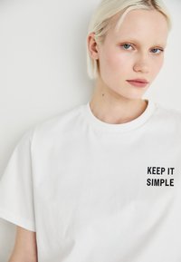 Hvit bomullst-skjorte med korte ermer og rund hals, med svart tekst "KEEP IT SIMPLE" trykket på venstre brystområde.