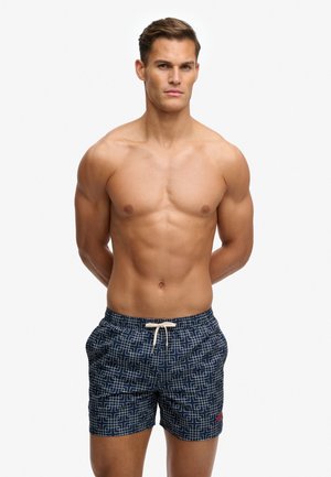 Muskularny mężczyzna stojący z rękami za plecami, ubrany w granatowe kąpielówki z białym geometrycznym wzorem i beżowym sznurkiem.