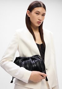 Bolsa de mão de couro preta plissada com fecho de cordão, segurada na mão, acompanhada por um blazer branco e blusa escura. Textura suave.