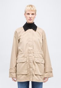 Άτομο με κοντά ξανθά μαλλιά φοράει μπεζ σακάκι με μαύρο γιακά και μεγάλες μπροστινές τσέπες πάνω από μαύρο ρούχο με ψηλό λαιμό και μπλε τζιν.