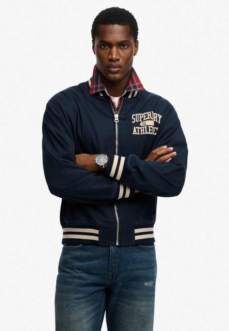 Uomo con le braccia incrociate che indossa una giacca bomber navy Superdry Athletics, camicia a quadri rossa, jeans blu e un orologio da polso su sfondo bianco.