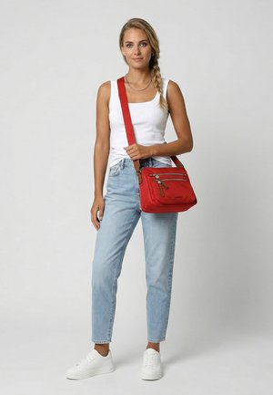 Blonde woman in a white tank top, light blue jeans, and white sneakers holding a red crossbody bag with multiple zippered pockets.