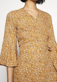 Vestido envolvente floral en mostaza con patrones en blanco y naranja. Cuenta con un escote en V y mangas acampanadas, fabricado en una tela ligera.