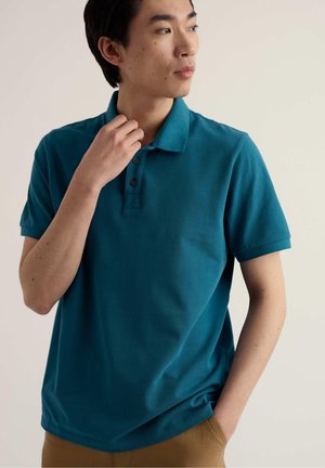 Junger Mann trägt ein türkisfarbenes Poloshirt mit kurzen Ärmeln und drei Knöpfen, schaut zur Seite, Hand in der Nähe des Kragens, vor einfarbigem Hintergrund.