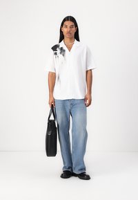 Camicia bianca a maniche corte con macchie di inchiostro nere, abbinata a jeans blu larghi e una borsa a tracolla nera. Tessuto liscio e vestibilità casual.
