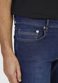 Jean en denim bleu foncé avec une texture lisse, comportant une poche à monnaie à l'avant, un bouton en métal argenté et des détails de couture en contraste.
