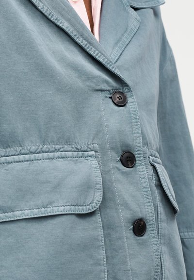 Gros plan sur une veste bleue avec des boutons noirs, de grandes poches à rabat et des coutures visibles, portée sur une chemise rose pâle.