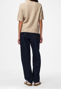 Personne portant un pull en maille beige à manches courtes, un jean large et foncé, et des baskets blanches à semelles marron, vue de dos.
