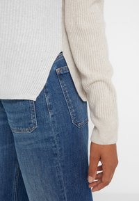 Beige geribde trui met mouwen en zijsplit, gedragen over blauwe denim jeans met achterzak, hand ontspannen langs de zijde.