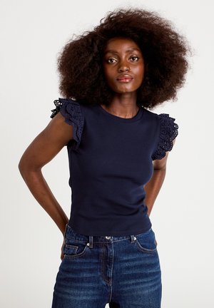 Woman with natural afro hairstyle wearing a navy blue top with eyelet lace ruffle sleeves and high-waisted blue jeans against a white background.