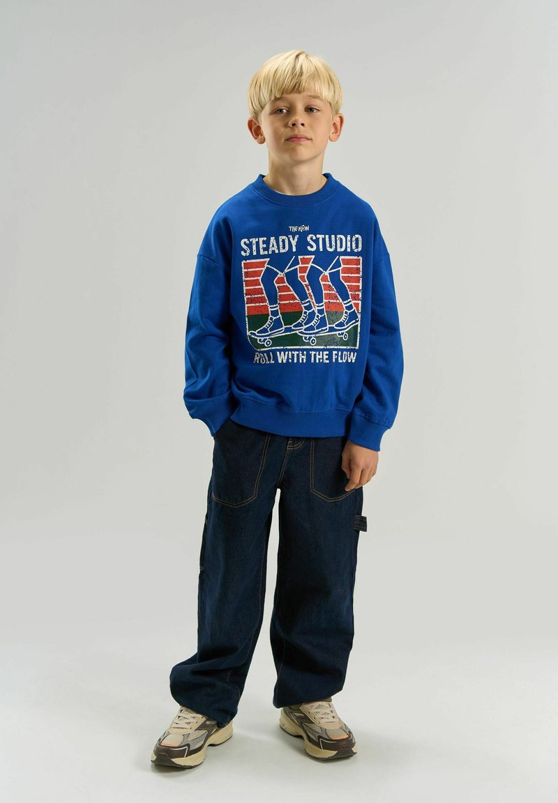 Garçon blond portant un sweat bleu avec un motif de skate et un jean large foncé, debout devant un fond uni.